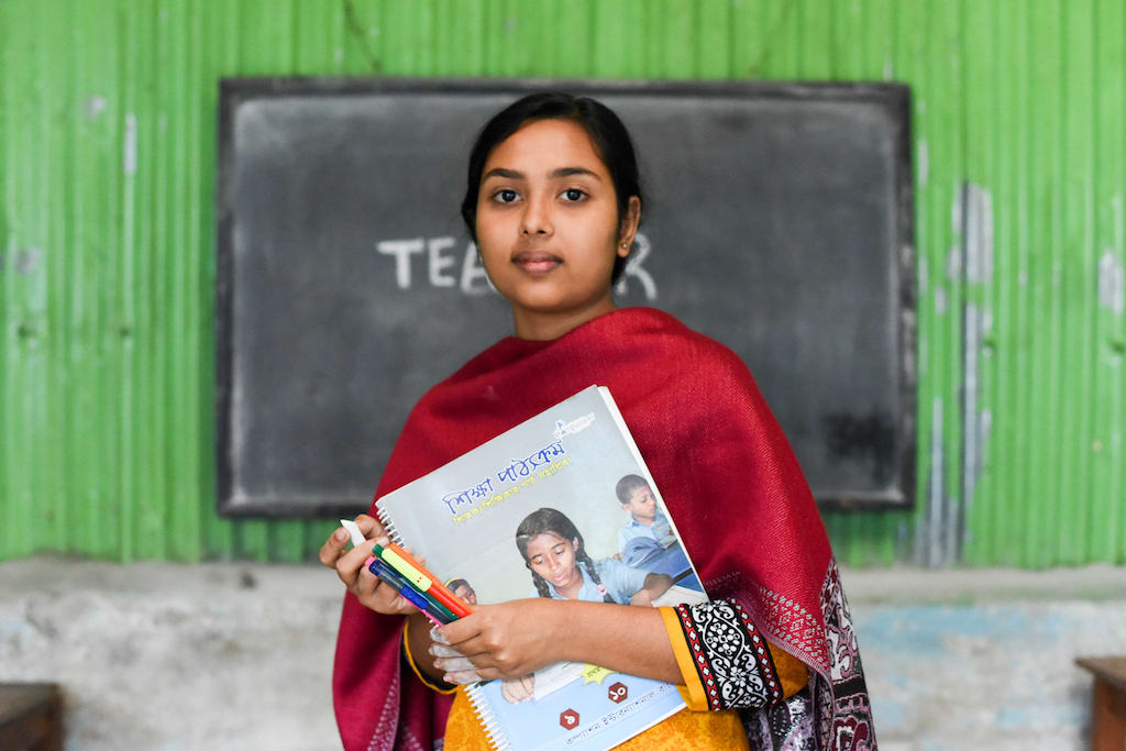 Teenage girl holds her workbook in her classroom, standing in front of a chalk board and a bright green wall.